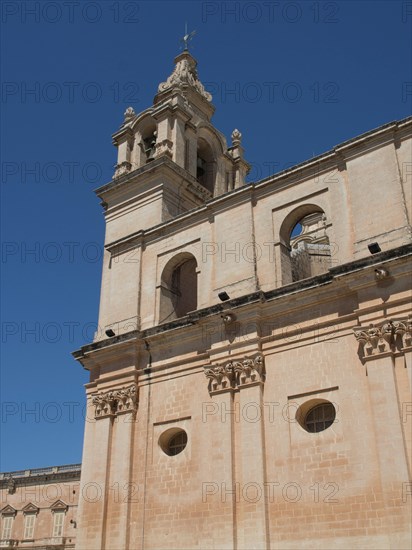 A tall church tower and sand-coloured buildings under a clear blue sky, the town of mdina on the