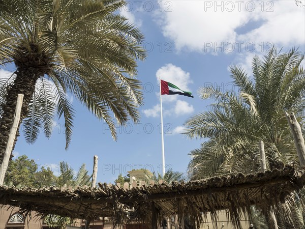 Flag of the United Arab Emirates waving in the wind, surrounded by palm trees and a thatched roof,