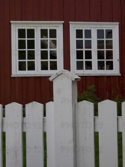 Red wooden house with two large white windows behind a willow fence, colourful wooden houses behind