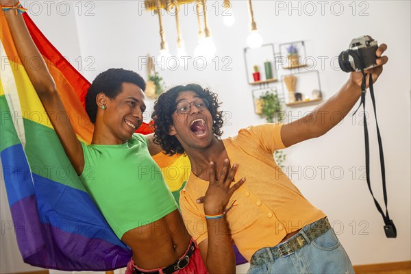 Gay latin young couple taking a selfie with a camera while raising rainbow lgbt flag at home