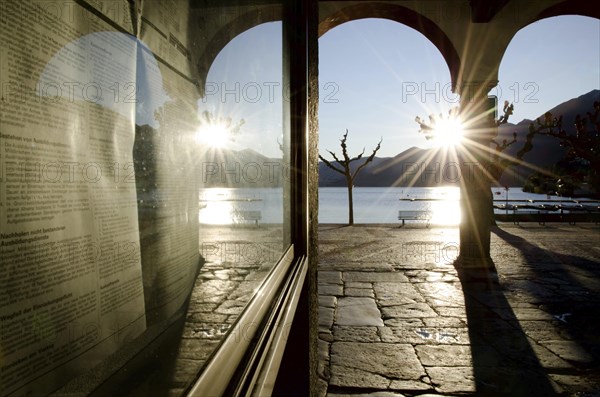 Alley with Arches on the Waterfront to Lake Maggiore with Mountain and Sunbeam and Reflection on a