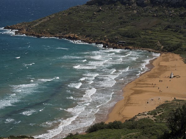 View of a quiet beach with golden sand and waves in the turquoise sea, the island of Gozo with