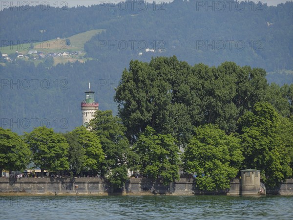A lighthouse behind trees on the shore of a lake, in the background mountains with forest, view of