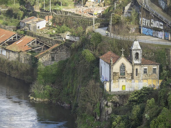 Small chapel and surrounding buildings on an overgrown, rocky cliff by a river with graffiti on