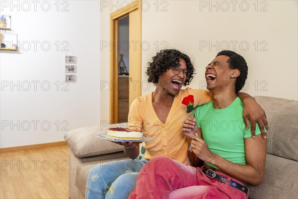 Gay couple embracing celebrating love with a rose and cake sitting together in the sofa at home