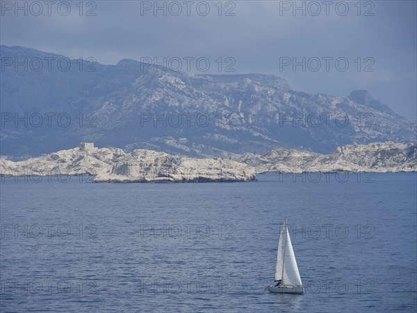 Sailboat sailing on a calm sea in front of a rocky mountain landscape and cloudy sky, Marseille on