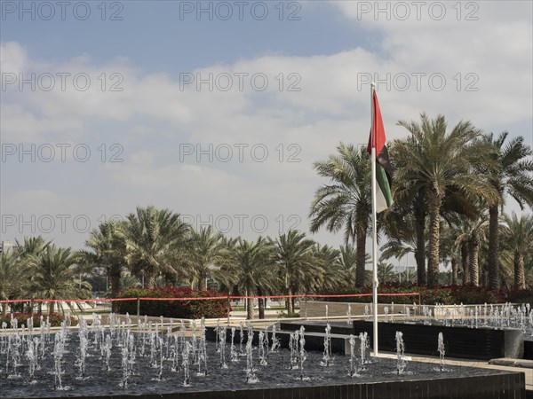 A fountain surrounded by palm trees under a slightly cloudy sky with a waving flag in the