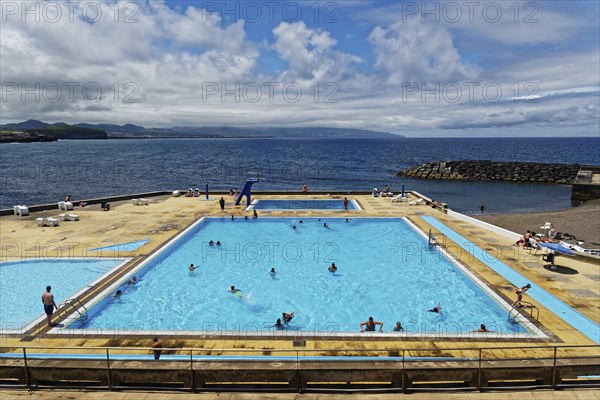 Large seawater swimming pool, filled with people, under a sunny, partly cloudy sky, coastal town of