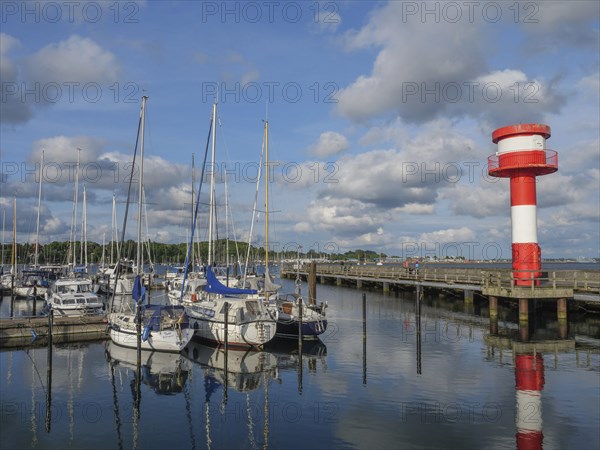 Clear harbour view with sailing boats and a red and white lighthouse under blue sky and scattered
