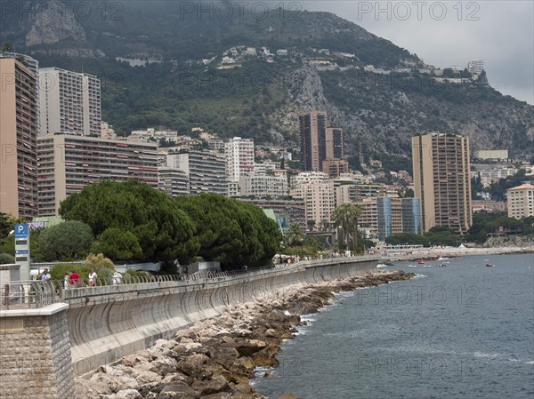 Coastline with promenade, skyscrapers and mountains behind, surrounded by sea and cloudy sky,