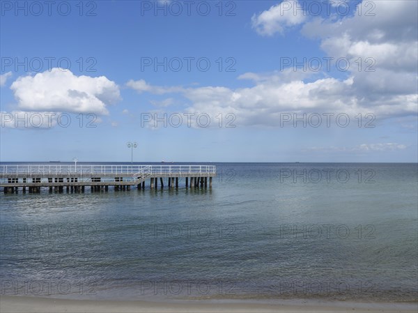 A jetty juts out over the calm waters of the sea, the sandy beach leads into the distance under a
