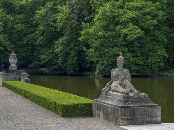 Stone statue in a park next to a lake, surrounded by dense green trees, historic castle with a