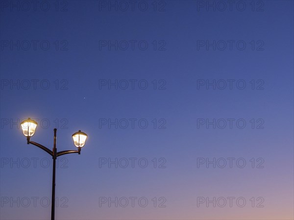 A street lamp shines against a dark blue sky at dusk, sunset in many colours on a beach with a calm