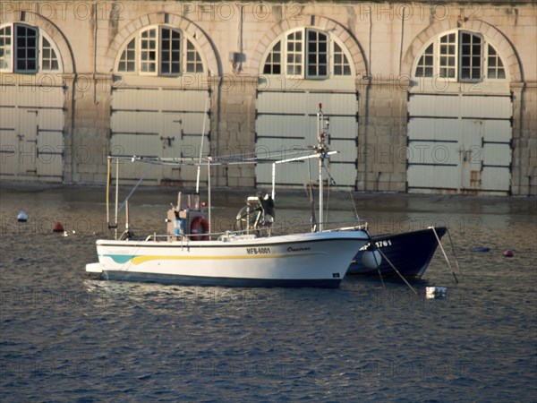 A boat in the harbour in front of a building with stone walls and many windows, small boats in the