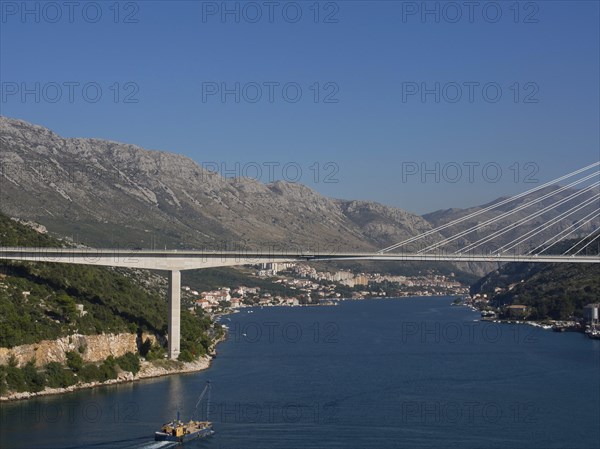 Wide view of a suspension bridge over a wide river, surrounded by a city and mountains under a