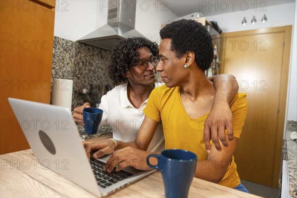 Romantic latin gay couple using laptop and drinking coffee sitting in a domestic kitchen at home