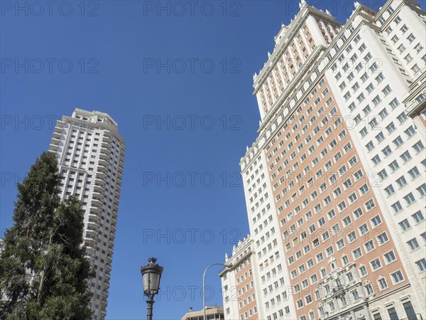 Two tall, modern buildings under a clear blue sky, a street lamp and a tree in the foreground,