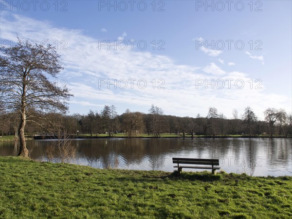 Park bench on the lakeshore with a wide view of the grassy banks and wooded background, small lake