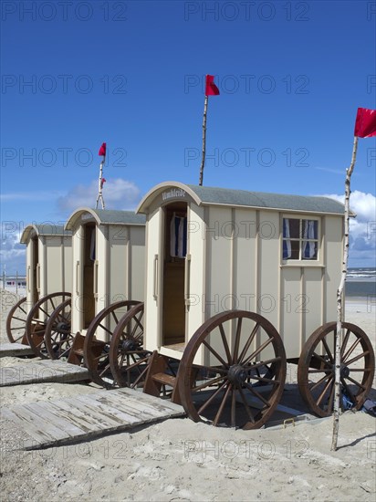 Three beige changing cabins with large wooden wheels on a beach under a blue sky with clouds,