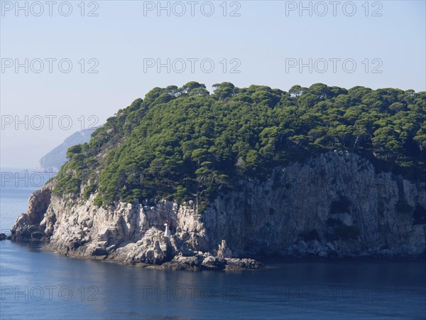 Rocky coast with densely overgrown trees and a small lighthouse, the sea calm and blue, the old