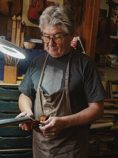 A luthier wearing glasses and an apron is intently working on a cello under a desk lamp