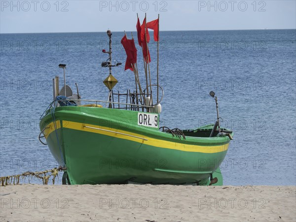 Green boat with red flags standing on sandy beach in front of calm sea under blue sky, green and