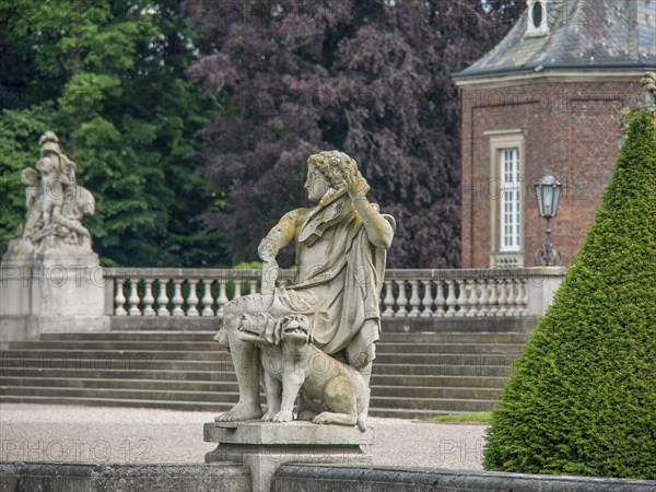 Stone sculpture in front of a castle with stone stairs and balustrades, surrounded by green bushes