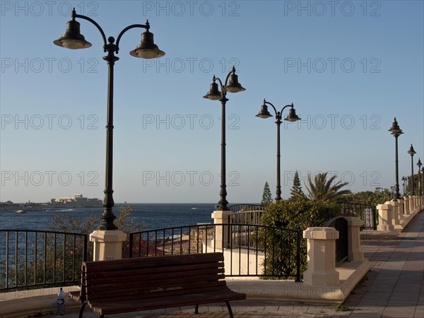 Promenade along the sea with benches, decorative lamps and palm trees, railings and clear sky,