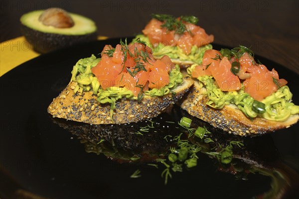 Gourmet bruschetta topped with avocado and salmon on a black plate, garnished with fresh dill