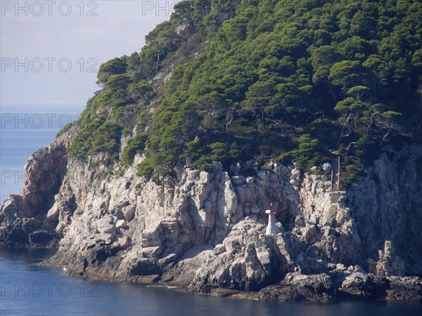 Rocky coast with densely overgrown trees and a small lighthouse overlooking the calm blue sea, the