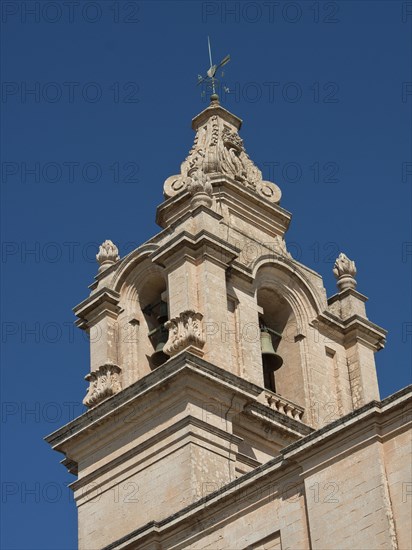 A large stone church tower with architectural details rising into the blue sky, the town of mdina