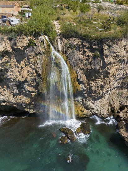 A waterfall cascades from a cliff into turquoise-coloured water in a rocky, natural setting, aerial