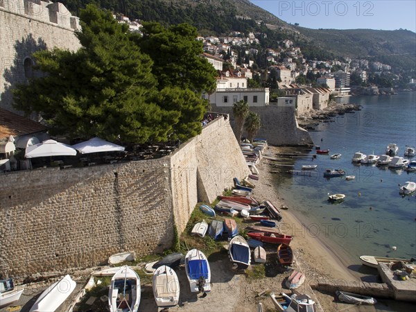 Boats moored on the shore next to a city wall, the sun shining brightly and the clear sky adding to