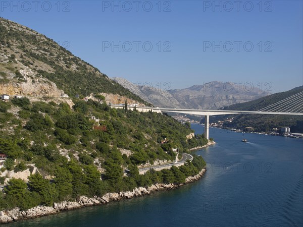 Landscape with coastal road and a bridge over a wide river, surrounded by green hills, the old town