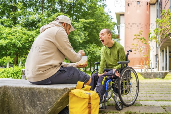 Male caucasian adult university student feeding a friend with cerebral palsy sitting on wheelchair