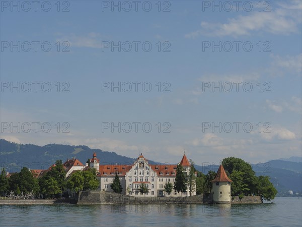 A castle with red roofs on a lake, surrounded by trees and mountains in the background, view of a