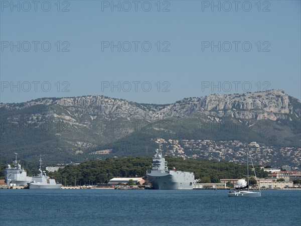 Several warships and a sailing boat in the harbour, green coast and mountain range in the