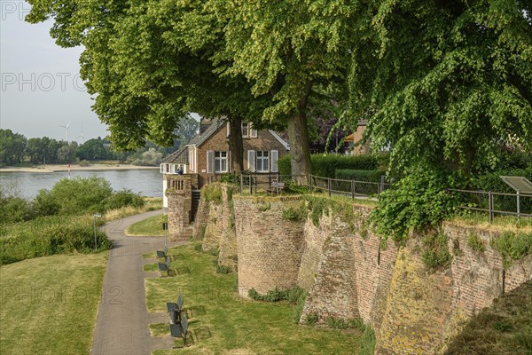 An old house on a river bank with garden and green trees, wall in the foreground, summer ambience,