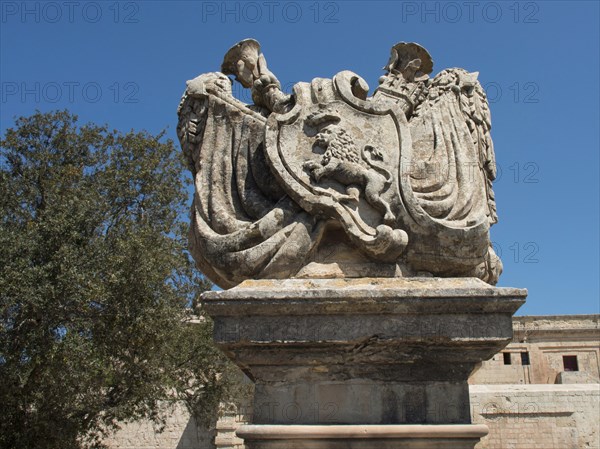 A large stone statue with coat of arms motifs in front of the old castle and trees in the