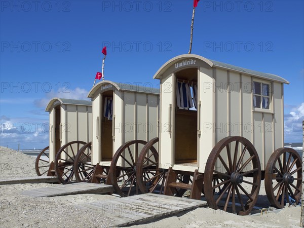 Beach with changing cabins and people on the shore, red flags and blue sky in the background,