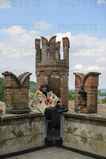 Contemplative older woman sits on a brick balcony overlooking a lush landscape, her expression