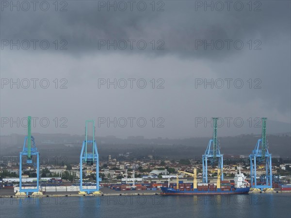 Large blue harbour cranes lifting containers, cloudy sky and a ship in the harbour industrial area