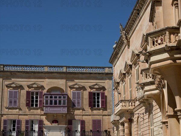 Two buildings with historic stone walls and shutters under a clear sky, the town of mdina on the