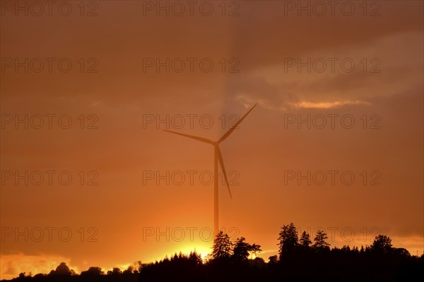 Silhouette of a wind turbine against an orange sunset sky with trees in the foreground, Upper