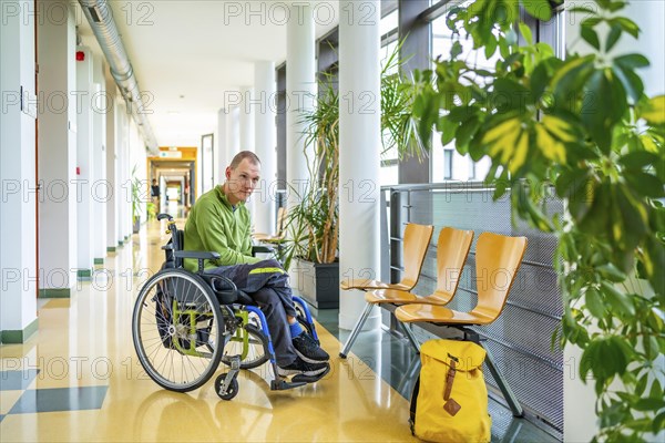 Man with cerebral palsy in wheelchair in the university waiting in the corridor next to school bag