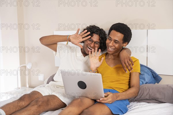 Gay couple waving during a video call with laptop lying on bed