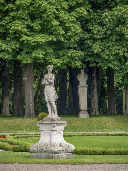 A statue in a green park with tall trees and another monument in the background, historic castle