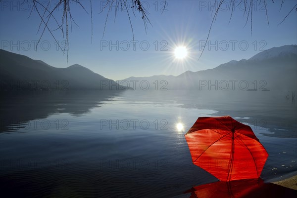 Red Umbrella Leaning on the Sand Beach on Alpine Lake Maggiore with Mountain and Sunbeam and