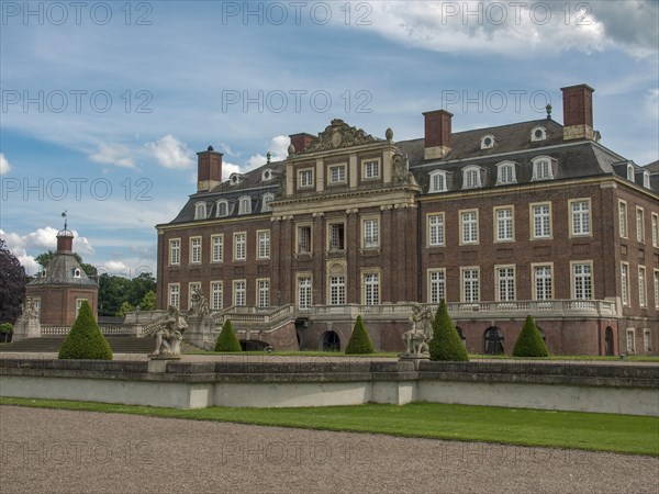 Historic castle with manicured lawn and sculptures, surrounded by a stone wall under a cloudy sky,