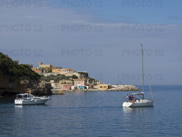 Sailing boats in front of a coastal landscape with cliffs and small houses under a partly cloudy